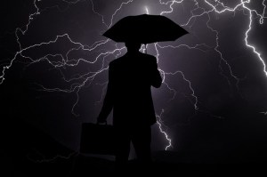 A silhouetted man holding up an umbrella as a lightning storm rages around him against a slate grey sky. He is holding a briefcase.