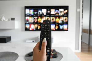 Woman's manicured hand pointing a remote control at a large screen television on the opposite wall in a spacious modern room with white minimal furnishing.
