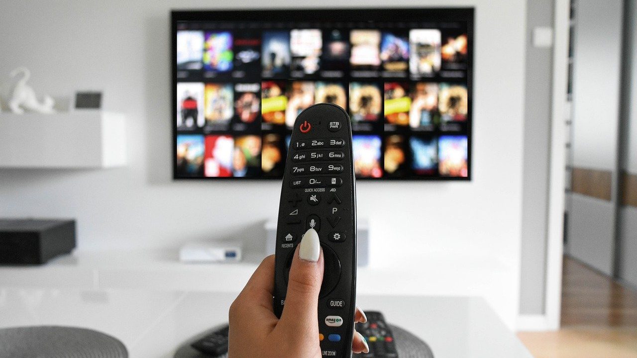 Woman's manicured hand pointing a remote control at a large screen television on the opposite wall in a spacious modern room with white minimal furnishing.